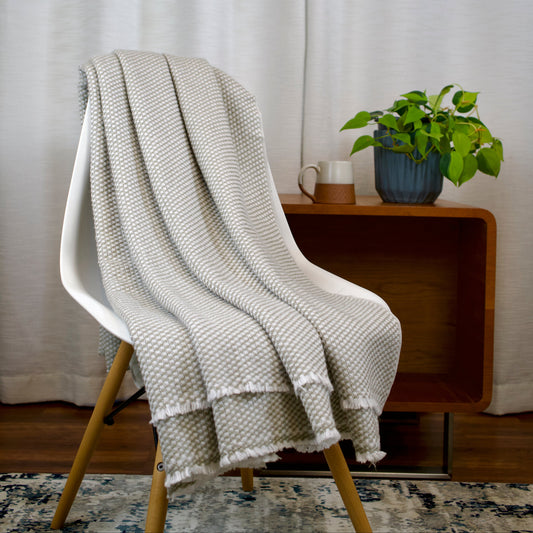 Light gray textured blanket draped over a white chair with a wooden side table and plant in the background.