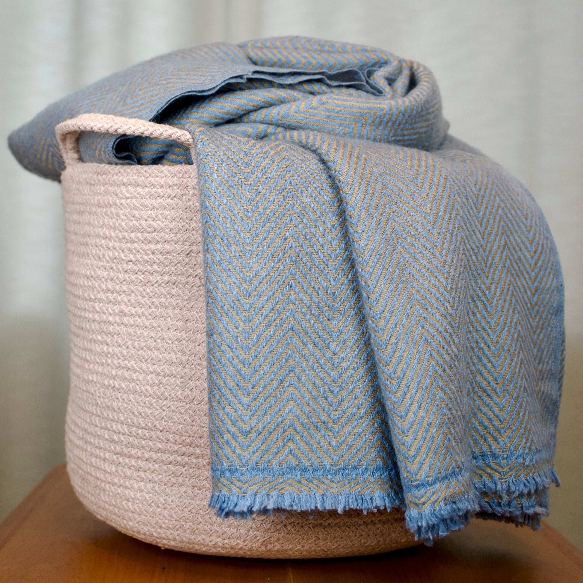 Blue and golden woven blanket draped over a beige woven basket on a wooden surface with a neutral background