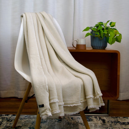 White cashmere blanket draped over a chair with a small table and plant in the background